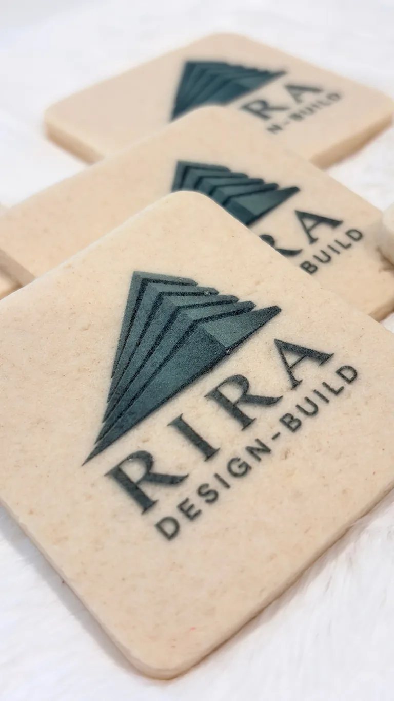 Three cream-colored wooden coasters with RTRA Design Build logo featuring a geometric arrow design in dark teal ink