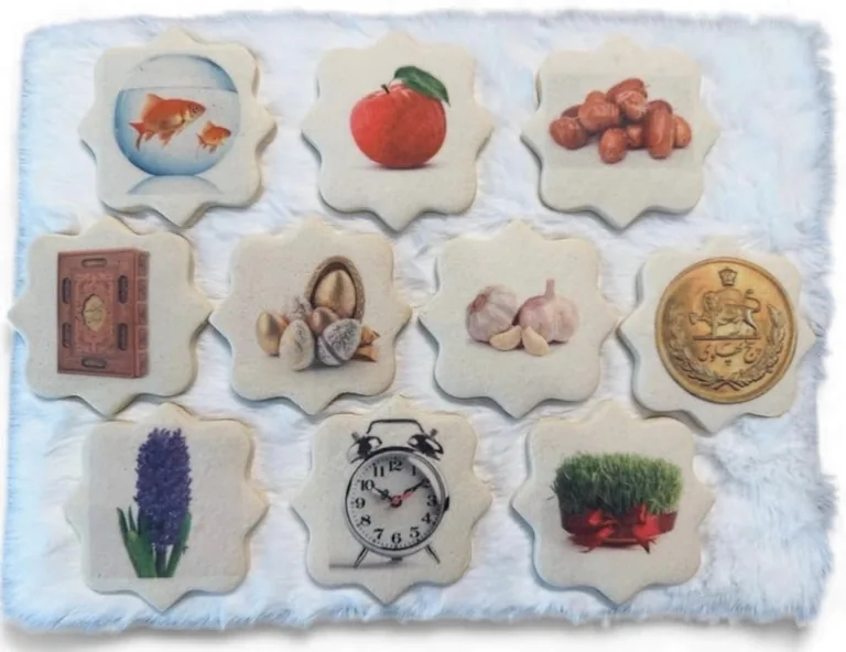 Collection of decorative ceramic tiles with various images including goldfish, apple, berries, book, flowers, garlic, alarm clock, coin, and lavender arranged on blue fabric